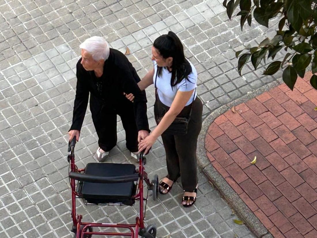 Cuidadora acompanyant una senyora gran que camina amb caminador pel carrer, agafant-la del braç